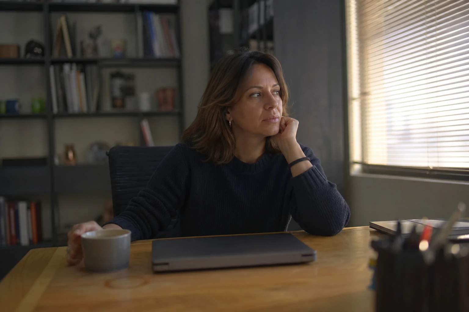 A small-business owner at an ordinary office desk in the late afternoon, laptop closed, wondering whether AI assistants name her brand when a customer asks a question