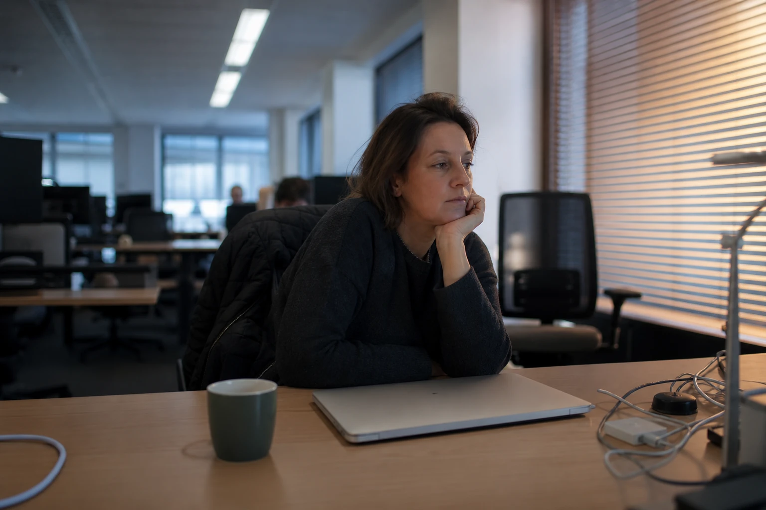 A marketing manager thinking at an ordinary office desk with a closed laptop in the late afternoon, wondering whether AI answers cite her brand
