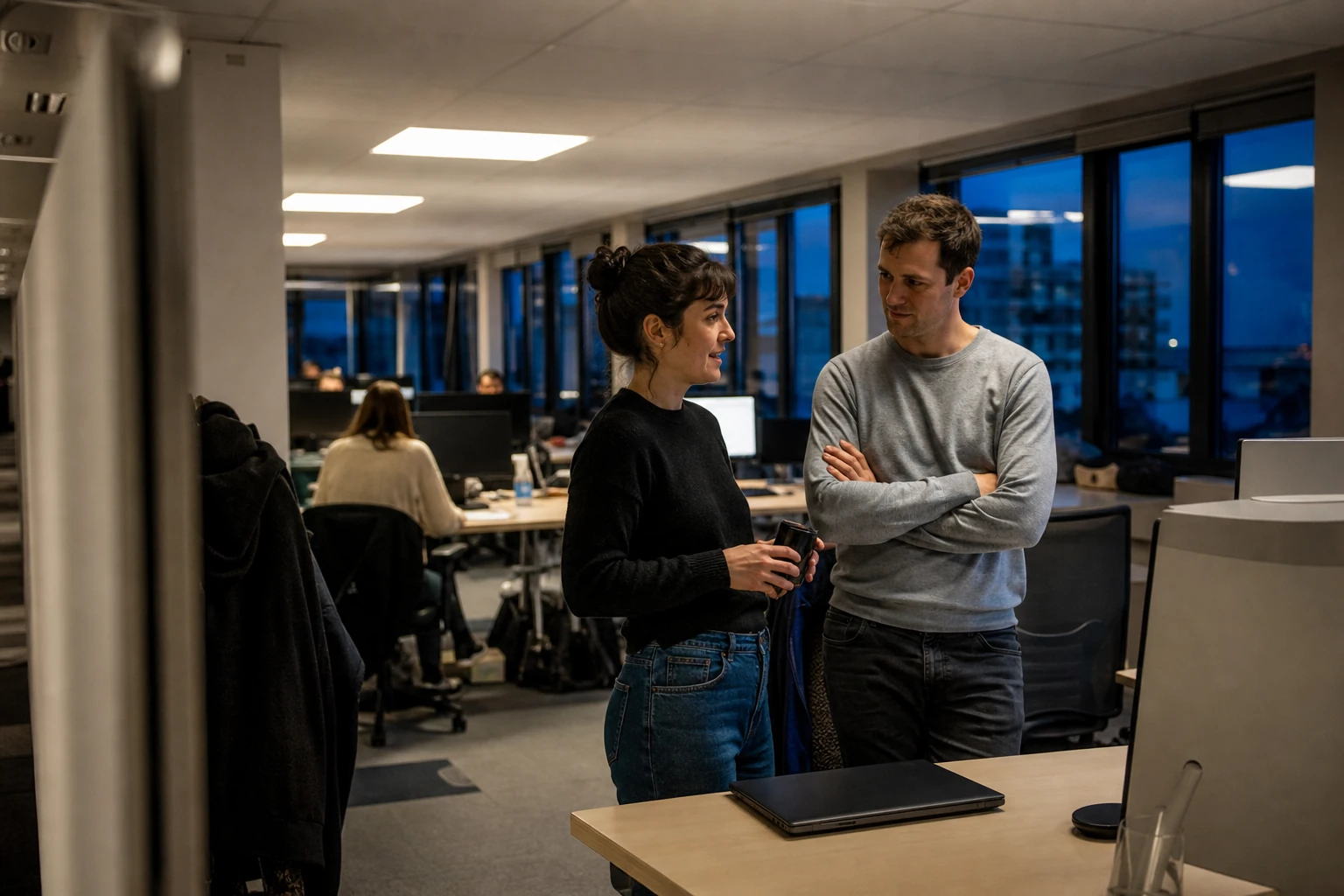 Two colleagues in an informal discussion near a desk in an open-plan office at the end of the day