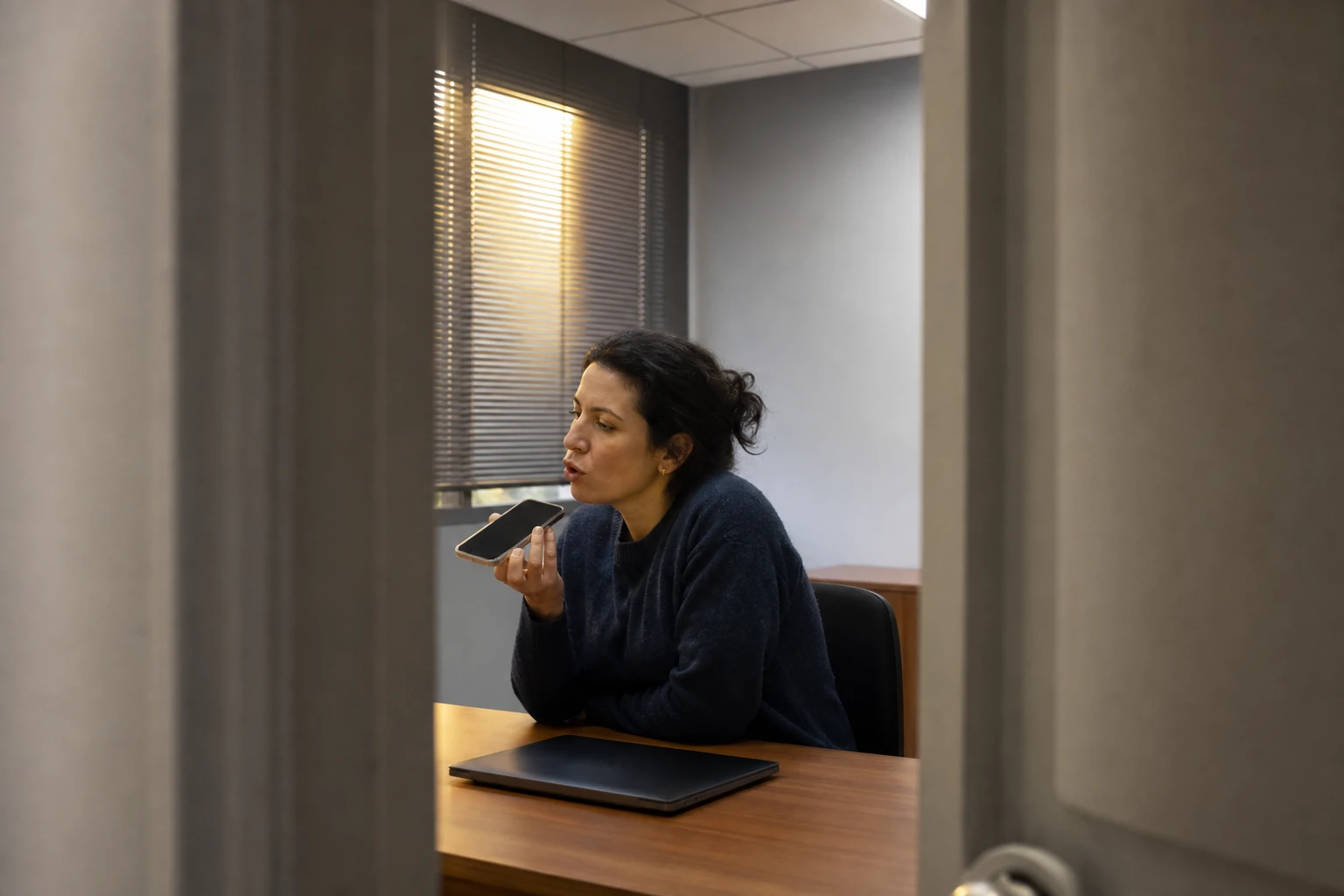 A person leaning on a desk speaking a question into a phone held near their face, a closed laptop beside them, waiting for a single spoken answer
