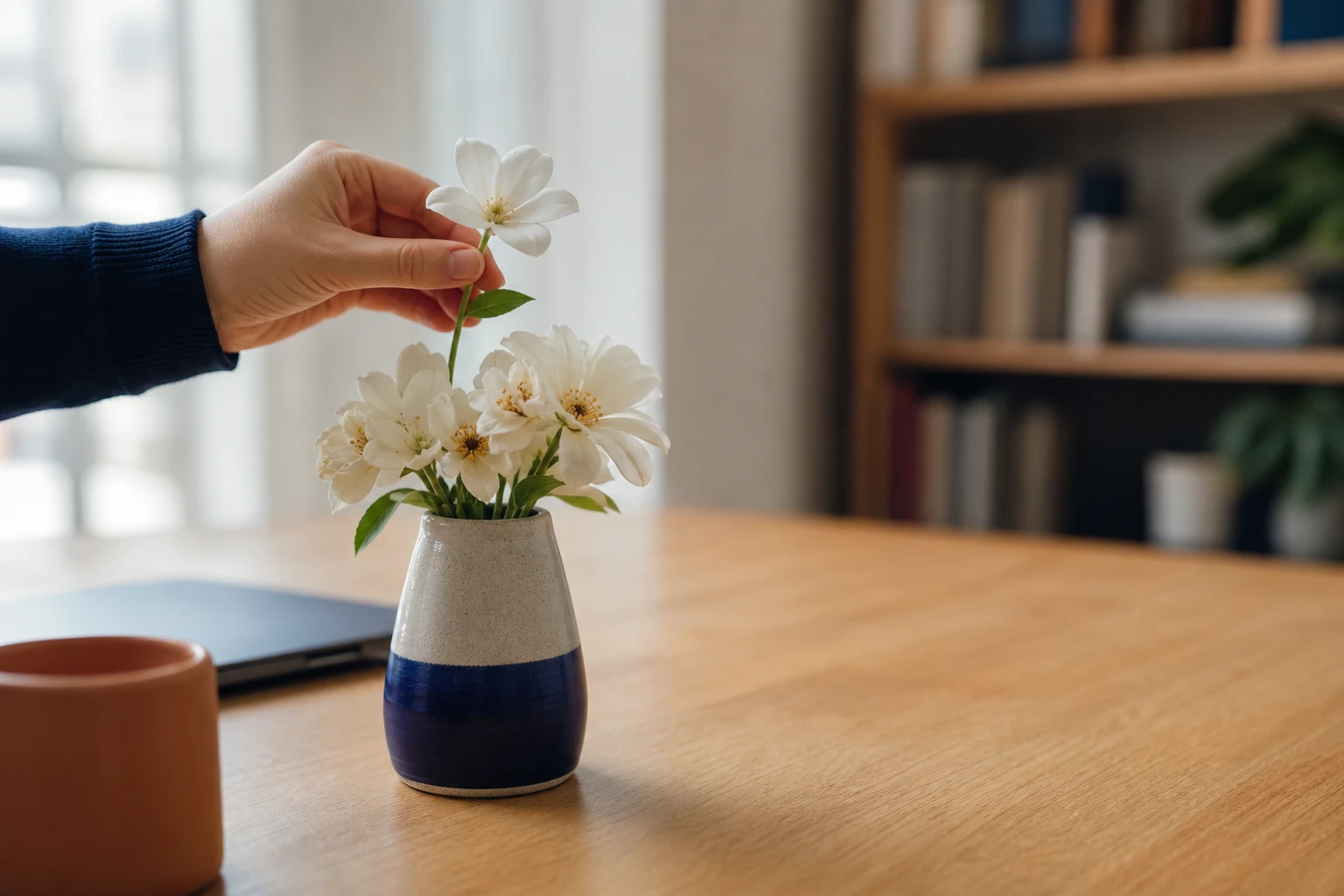 A hand placing one flawless artificial white flower into a ceramic vase of real fresh white flowers on a wooden desk, a closed laptop and a terracotta mug blurred beside them